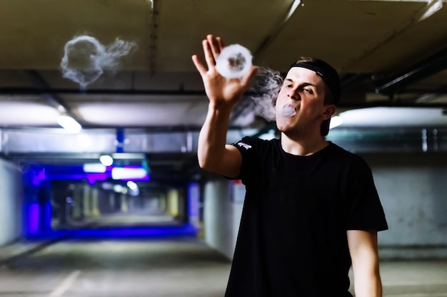 Man in cap smoke an electronic cigarette and releases clouds of vapor