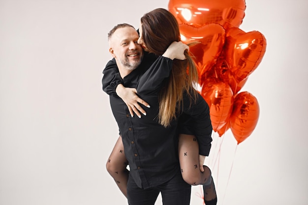 Man carrying woman piggyback while posing in studio near bunch of heartshaped red ballons