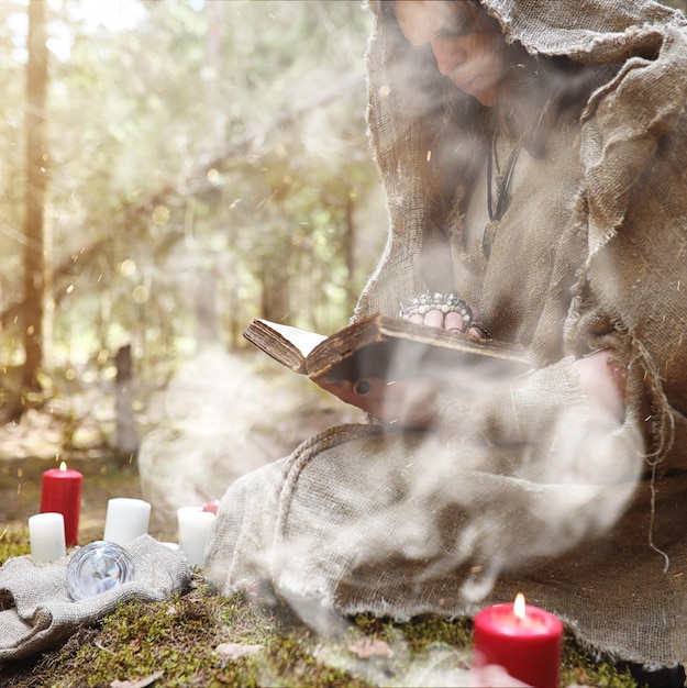 A man in a cassock spends a ritual in a dark forest with a crystal ball and bookxAxA