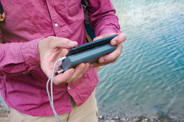 Man charges a smartphone with a portable charger. A man with a power bank in his hand on the background of the sea on a bright sunny day with clouds.