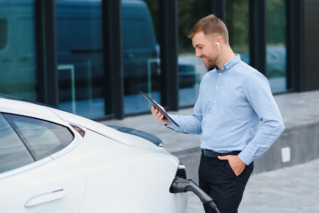 Man charging his electric car at charge station and using smartphone