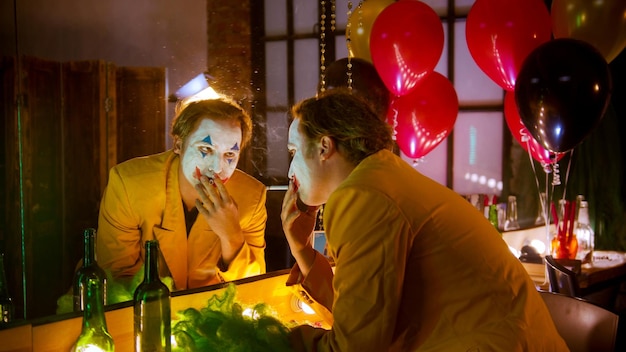 A man clown sitting in front of the mirror and smoking a cigarette