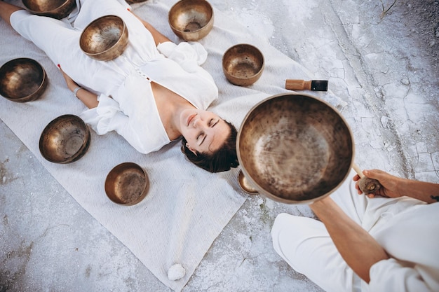 Photo man conducts therapy tibetan singing bowls for a girl lying on the ground in the desert surrounded by copper bowls