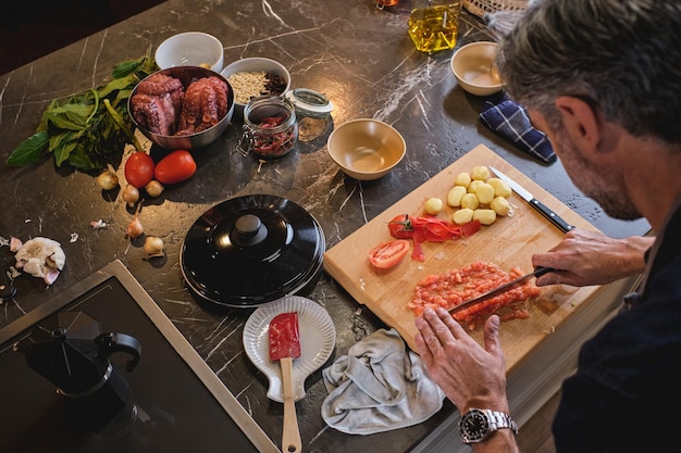 Man cutting pieces of tomato finely on wooden chopping board lying on kitchen counter full of various ingredients