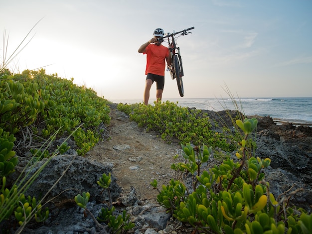 Man cyclist carries a bicycle in his hands, at ocean shoreline. Rocky beach.