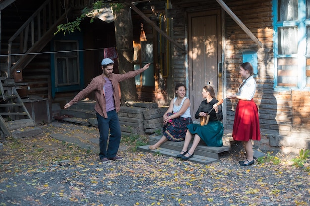 A man dancing russian dance women sitting on the stairs near the country house and talking to each other a woman playing balalaika