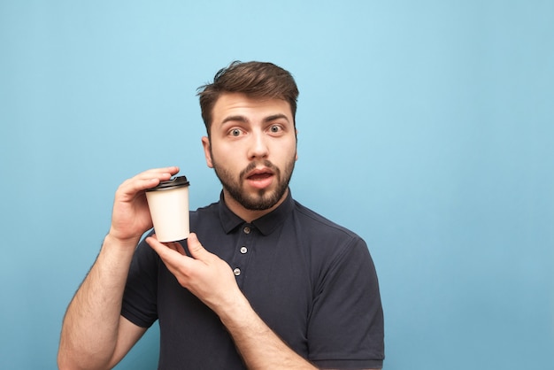 man in a dark shirt and beard, holding a paper cup of coffee in his hands on blue with an emotional face