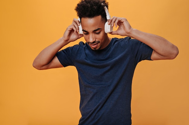 Man in dark Tshirt wears white headphones Snapshot of young guy in blue tee listening to music on orange isolated background