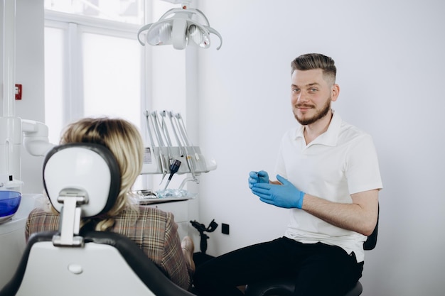 Man dentist in face mask and glasses doing treatment for patient blonde lady holding dental tools wearing rubber gloves Stomatology dentistry modern dental clinic concept