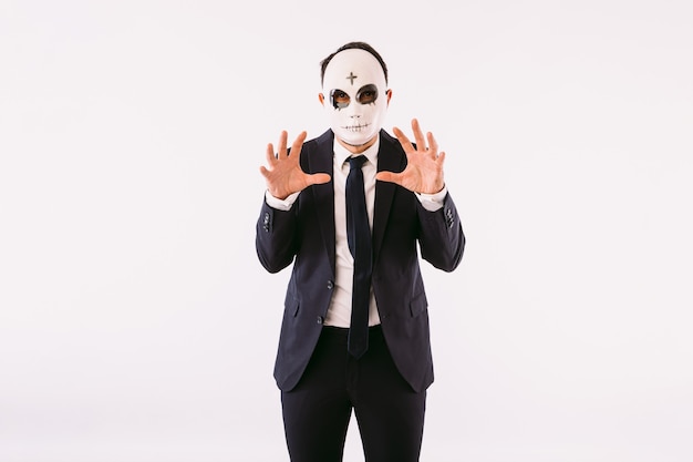 Photo man dressed in a suit and tie, wearing a killer's mask with a cross on his forehead for halloween, scaring with his hands. carnival and halloween celebration