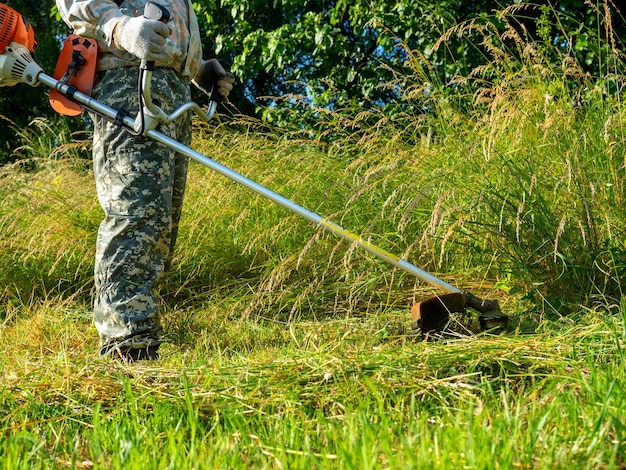 A man dressed in work clothes using a gasoline mower cuts long grass Summer rural area maintenance of the territory