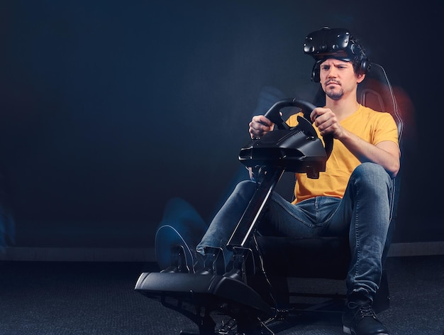 Man dressed in yellow shirt and jeans wearing VR headset driving on car racing simulator cockpit with seat and wheel.