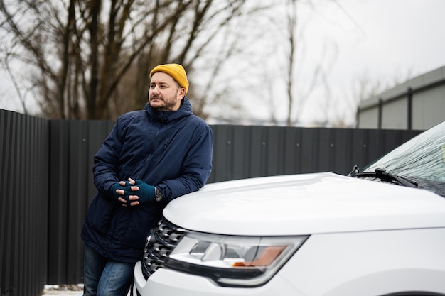 Man driver wear jacket and yellow hat against leaned on the hood of american SUV car in cold weather