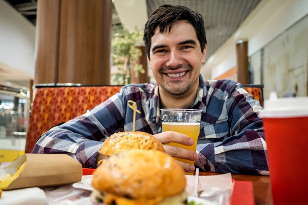 Man eating fast food burger and drinking beer alone in the open area of a restaurant in a shopping mall.