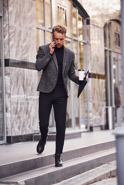 Man in elegant formal wear with cup of drink and notepad in hands is outside against modern building have a conversation by the phone.