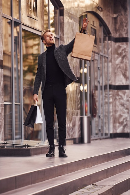 Man in elegant formal wear and with shopping packages is outside against modern building.