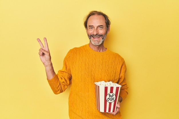 Man enjoying popcorn in yellow studio joyful and carefree showing a peace symbol with fingers