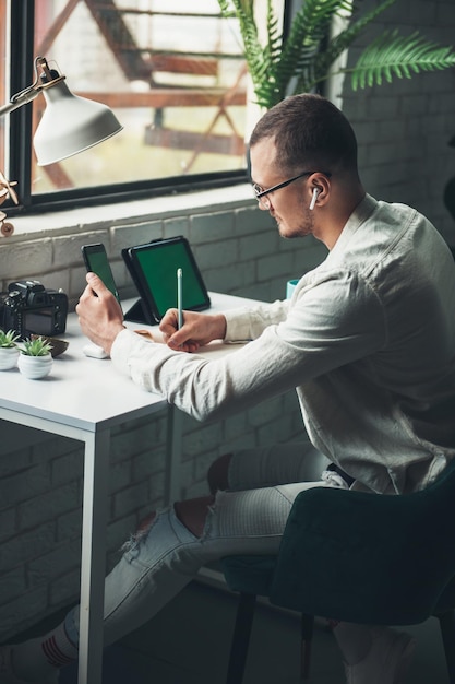 Man entrepreneur in eyeglasses sitting and working at table writing in planner holding smartphone an