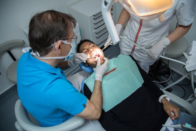 Man examining oral cavity of young African-American man working in in dental clinic with assistant.