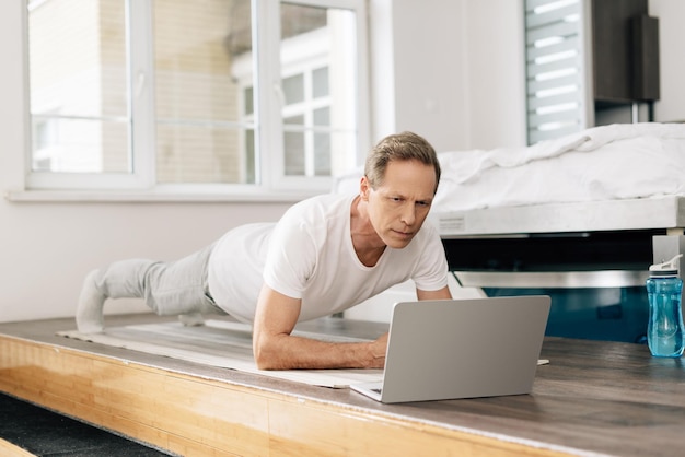 Man exercising on fitness mat while watching online working out on laptop