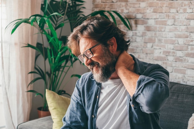 Photo man in eyeglasses holding his painful neck sitting on sofa at home caucasian guy suffering from neckache due to incorrect posture or joint inflammation frustrated mature male in severe pain