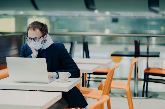 Man freelancer wears face mask to prevent spread of coronavirus and coughing uses laptop computer for distant work poses in cafe with cup of coffee reads information about symptoms of disease