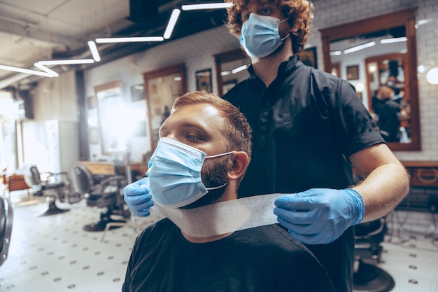 Man getting hair cut at the barbershop wearing mask during coronavirus pandemic. Professional barber wearing gloves. Covid-19, beauty, selfcare, style, healthcare and medicine concept.