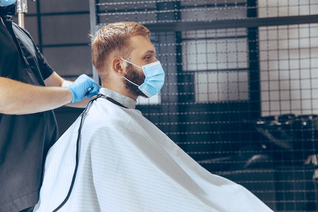 Man getting hair cut at the barbershop wearing mask during coronavirus pandemic. Professional barber wearing gloves. Covid-19, beauty, selfcare, style, healthcare and medicine concept.