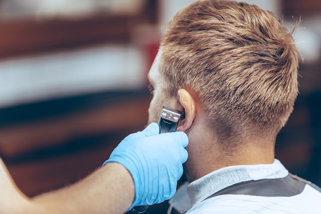 Man getting hair cut at the barbershop wearing mask during coronavirus pandemic. Professional barber wearing gloves. Covid-19, beauty, selfcare, style, healthcare and medicine concept.