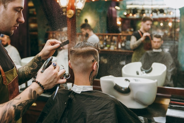 Man getting trendy haircut at barber shop. The male hairstylist in tattoos serving client.