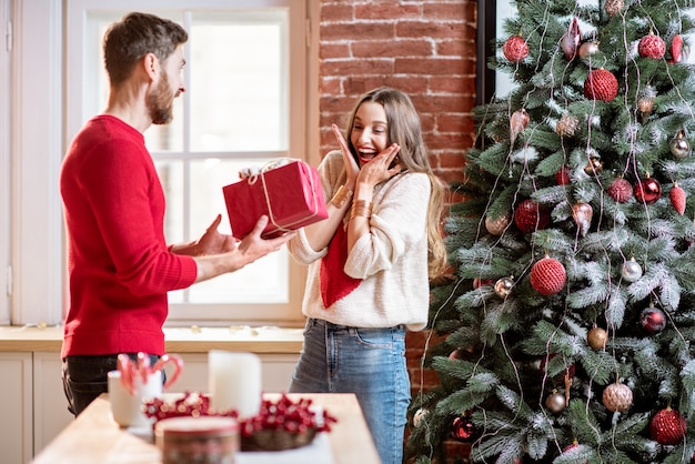 Man giving a New Year gift for a young excited woman standing near the Christmas tree at home