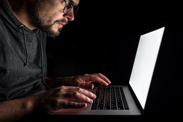 A man in glasses looks seriously at a laptop screen, works at a computer at night in the dark.