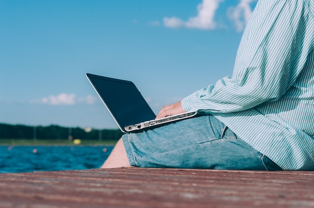 A man in glasses and a shirt working on a laptop in the fresh air, against the space of a river.