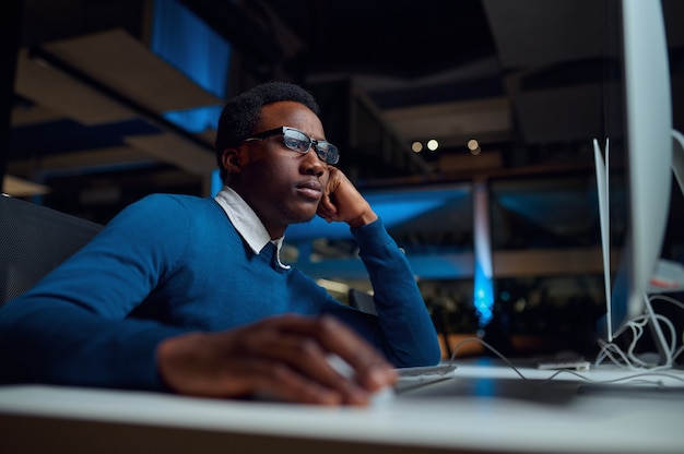 Man in glasses works on computer, office lifestyle. Male person at desktop, dark interior, modern workplace