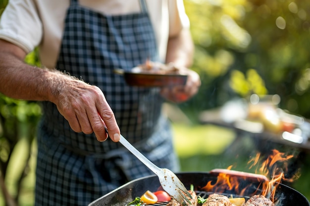 Man Grilling Vegetables and Meat Outdoors in Sunlit Garden Wearing Checkered Apron