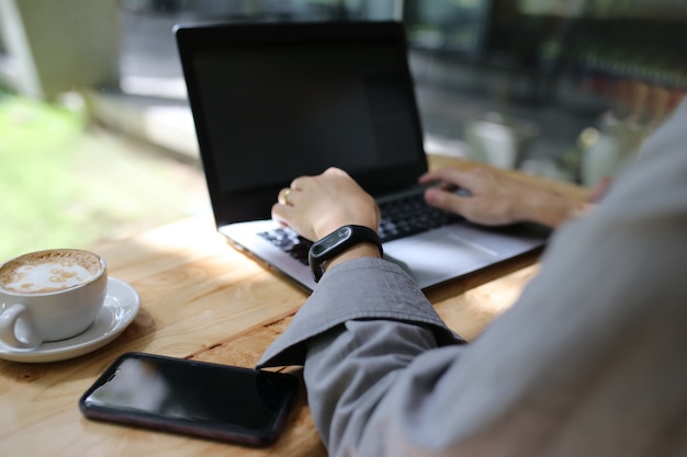 man hand and finger typing laptop keyboard on wooden table with phone and coffee cup, smart watch