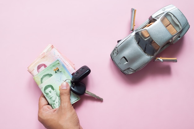 Man hand holding car key and Thai banknotes with car model on pink background