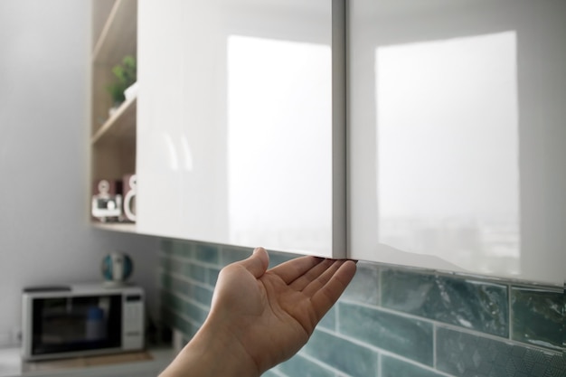 Man hand open the white cupboard cabinet door on a background of blue kitchen interior. A modern kitchen cabinet made without handles with an additional small opening for opening the door