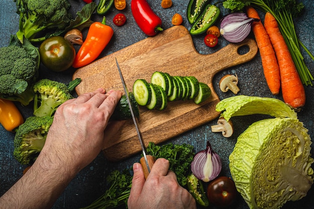 Man hands chopping cucumber on wooden cutting board with knife and assorted fresh vegetables