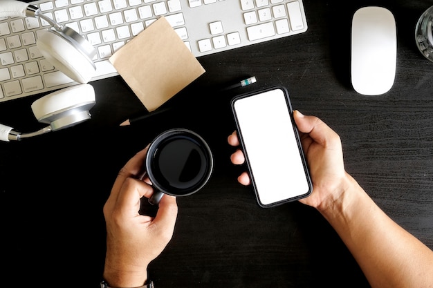 Man hands holding coffee mug and mobile phone on dark office desk.