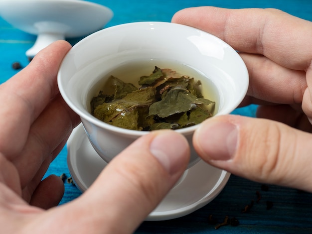 In man hands is a white porcelain bowl with fresh tea on a blue wooden background.