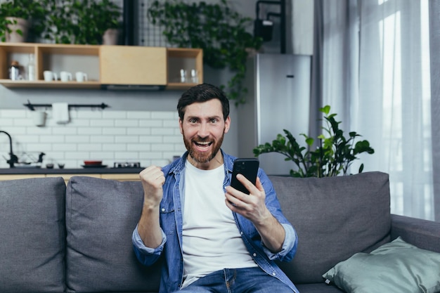 Man happy and shocked by the good news looks at the camera and smiles holding a phone in his hands sitting on the couch at home