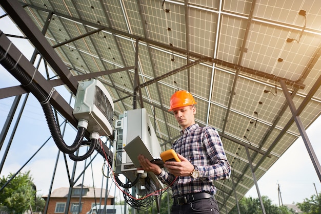 Man in hardhat standing with laptop near electrical equipment in a solar power station