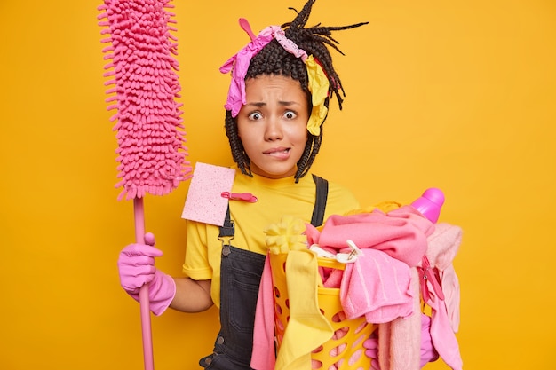 man has nervous expression bites lips holds mop and laundry basket with dirty iems dressed in casual onalls poses on yellow 