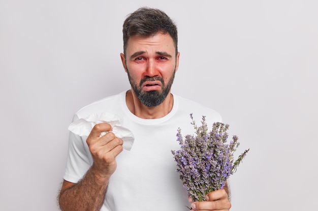  man has runny nose holds handkerchief cannot stop sneezing has allergy to lavender wears casual t shirt red itchy eyes and nose isolated on white wall. Allergic reaction