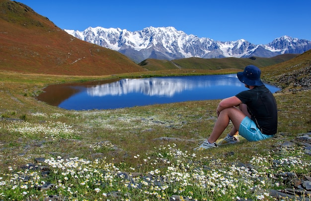 A man in a hat is resting in a flower meadow near a lake overlooking the snowcapped mountains in georgia svaneti