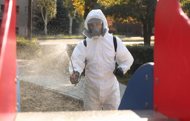 Man in hazmat suit spraying disinfectant onto slide at children's playground Surface treatment during coronavirus pandemic