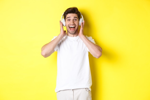 Man in headphones looking surprised and happy, listening awesome song, standing over yellow wall