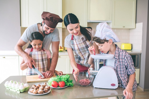 Man helps her daughter to cut meat with knife. Son is putting piece of it into meat grinder. Mom looks at it and holds bowl with both hands.