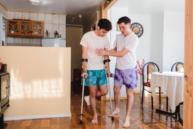 Man helps his gay partner who is using crutches inside their house LGBT couples cohabitation routin